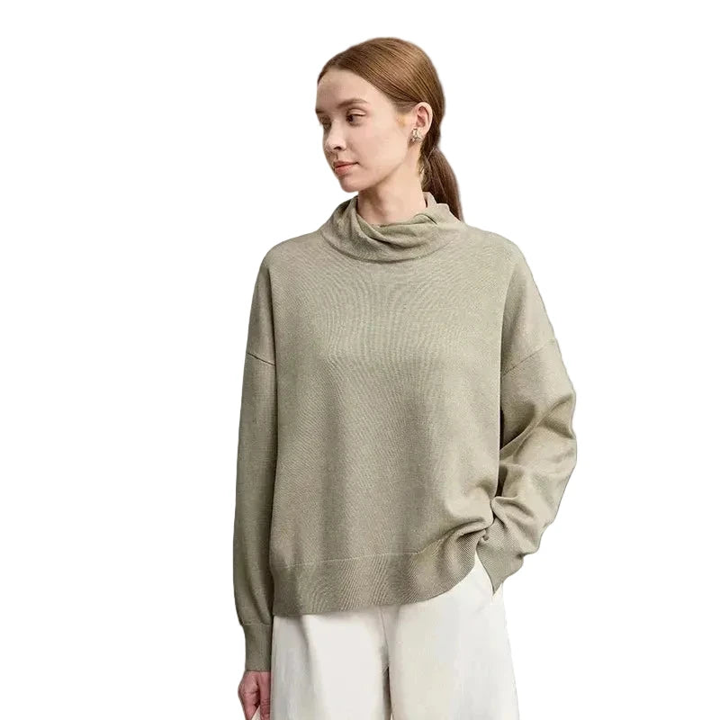 A woman wearing a light sage green ribbed knit mock-neck sweater and white pants, standing against a neutral light gray background. The brand name 'AMII' is printed in black in the top-left corner.
