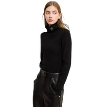 A woman modeling a black AMII turtleneck sweater with a small silver-toned decorative accent at the collar, paired with black pants, against a plain off-white background.