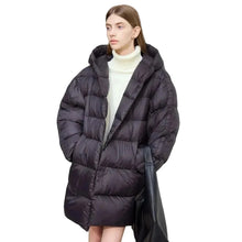 A woman wearing a long black puffer coat with a high collar over a white turtleneck sweater, standing against a plain light gray wall. The AMII brand logo is visible in the top left corner.