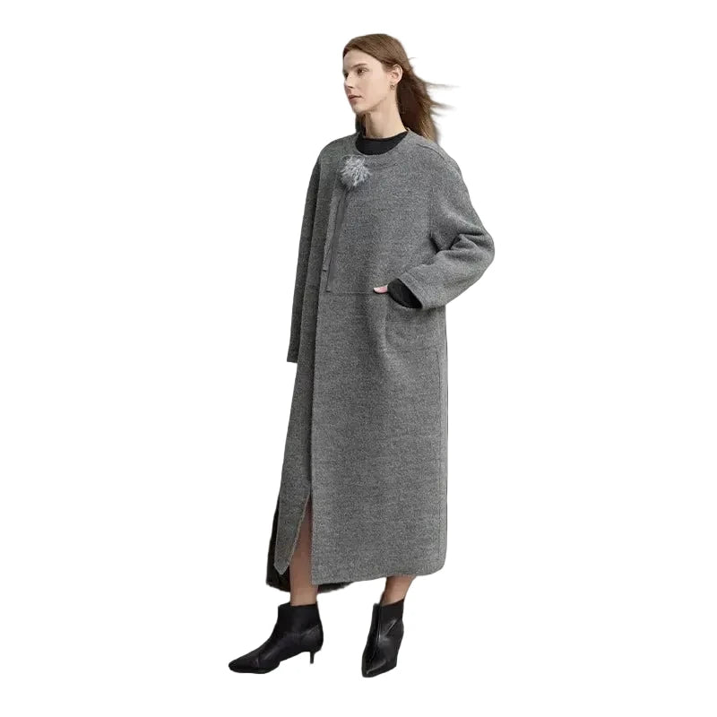 A woman models a long gray knit coat with a black crew neck underneath, standing against a minimalist light gray studio wall. The brand name 'AMII' is printed in black in the top-left corner.