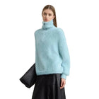 A model wearing a light blue fuzzy turtleneck sweater and a black skirt, holding a black clutch bag against a plain light gray wall. The brand name 'AMII' is printed in black in the top left corner.