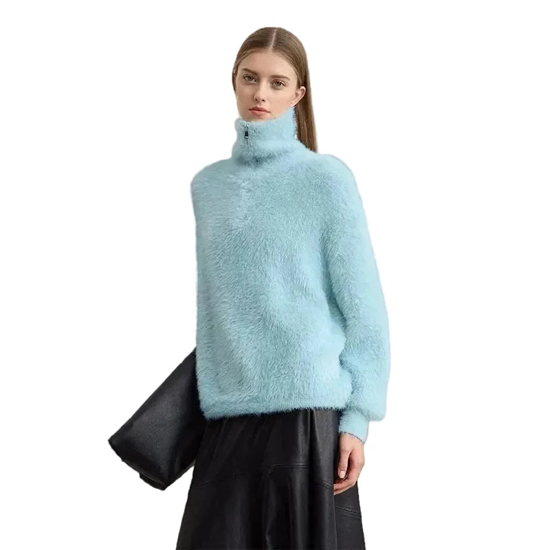 A model wearing a light blue fuzzy turtleneck sweater and a black skirt, holding a black clutch bag against a plain light gray wall. The brand name 'AMII' is printed in black in the top left corner.