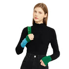 A woman modeling a black mock-neck long-sleeve knit top with half-finger gloves featuring green and light blue cuffs, holding a green shoulder bag strap against a plain off-white background. The brand name AMII is printed in the top left corner.